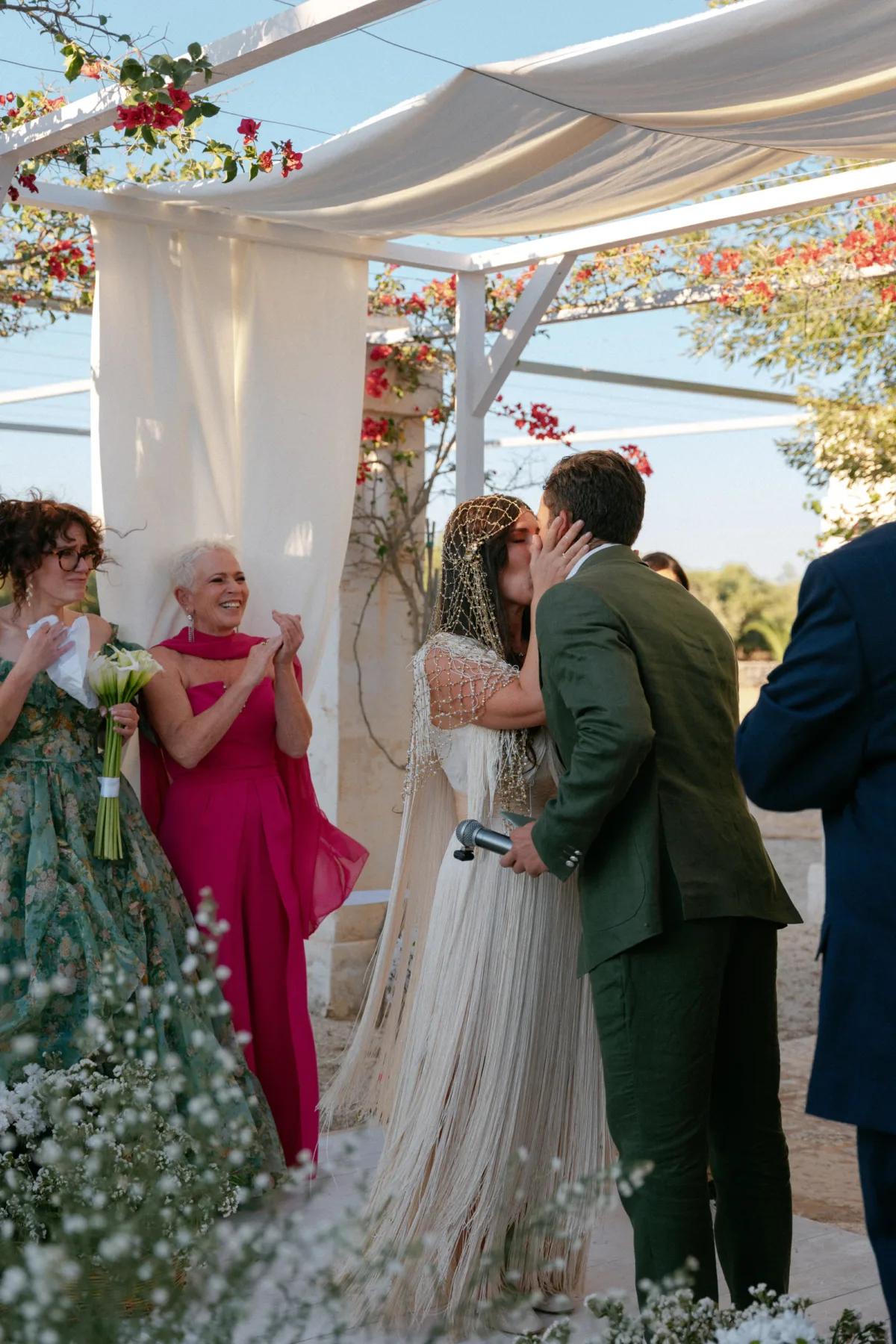 Multiple Day Destination Wedding in Masseria Potenti Puglia Italy 84 - Yael Wore Chana Marelus & A Handmade Beaded Veil for her Relaxed Italian Masseria Wedding Bride in a beaded veil holding her groom's face in her hands during the wedding ceremony as she kisses him. Puglia Italy outdoor wedding in the sunshine.