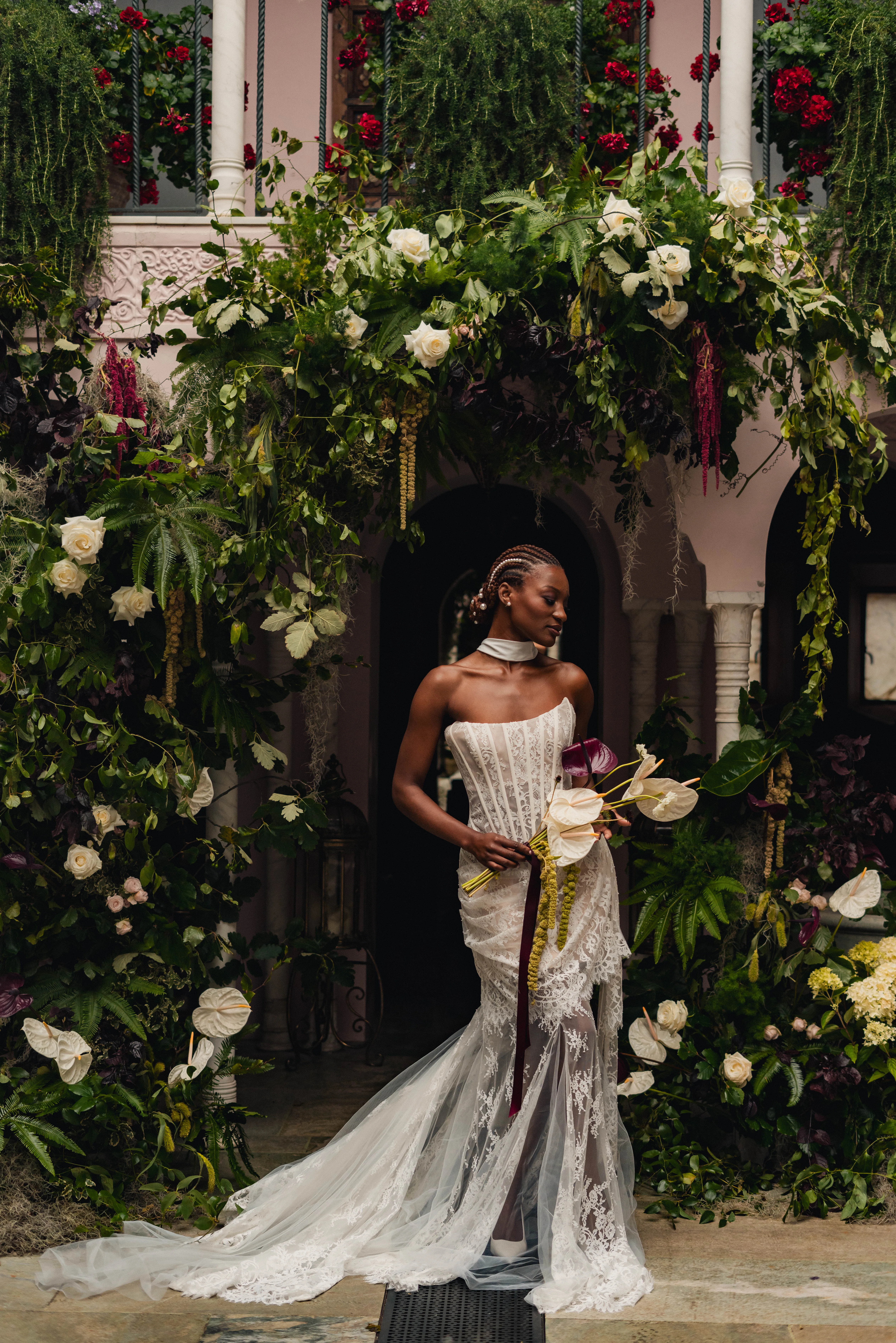 SS Port Lympne Safari Park New Orleans Wedding Editorial The Chamberlins Film Medium Format Photography London Surrey 250 - Wedding Inspiration That Celebrates The Joy, Diversity & Vibrancy of New Orleans Black model bride in a scene inspired by New Orleans, carrying a bouquet of orchids and surrounded by a broken arch of lush greenery at Port Lympne Resort wedding venue in Kent. She wears pearls in her braided hair.
