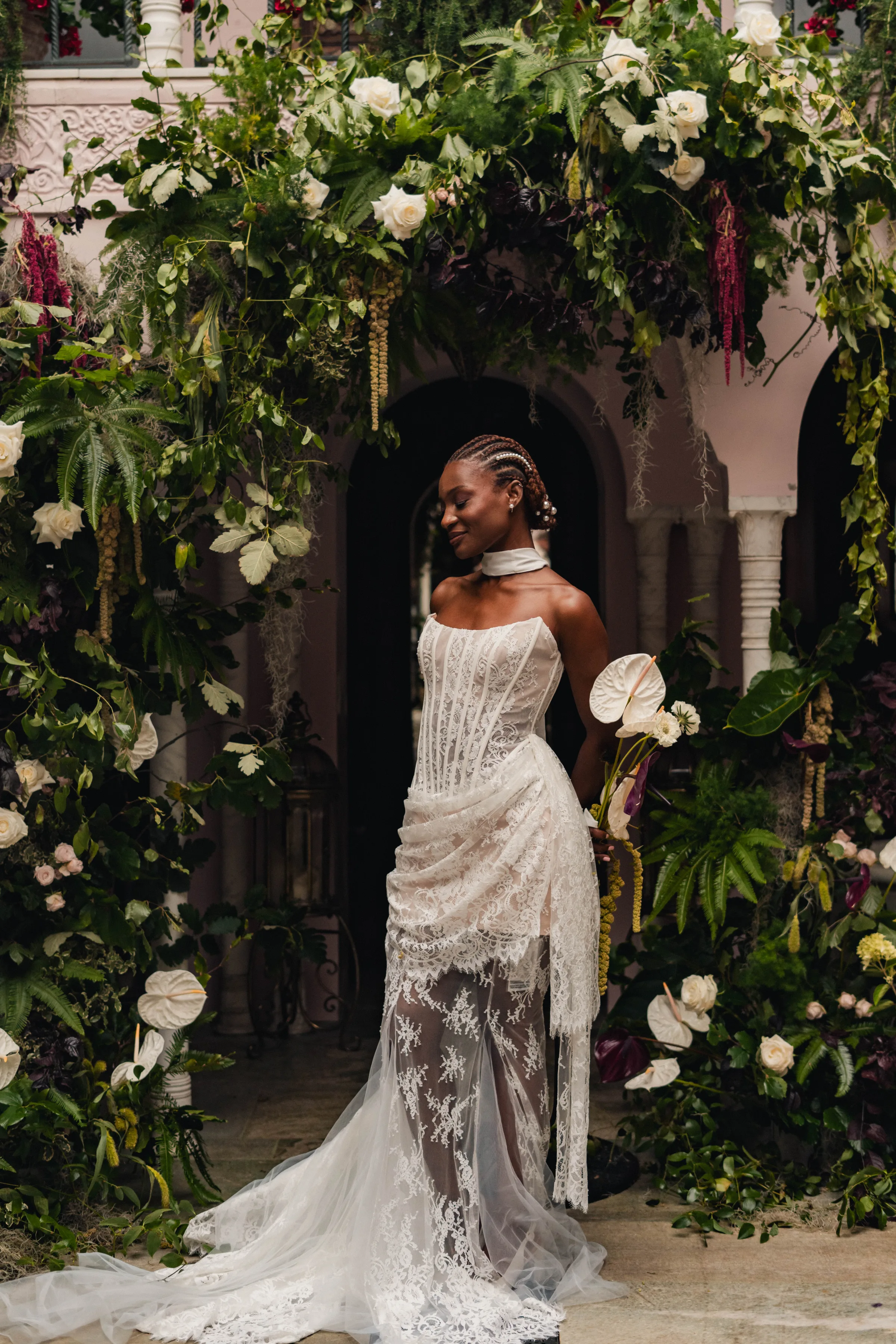 SS Port Lympne Safari Park New Orleans Wedding Editorial The Chamberlins Film Medium Format Photography London Surrey 257 - Wedding Inspiration That Celebrates The Joy, Diversity & Vibrancy of New Orleans Black model bride standing by a lush green broken arch featuring greenery, white roses and white orchids.. She carries a bouquet with large white orchids and wears pearls in her braided hair.