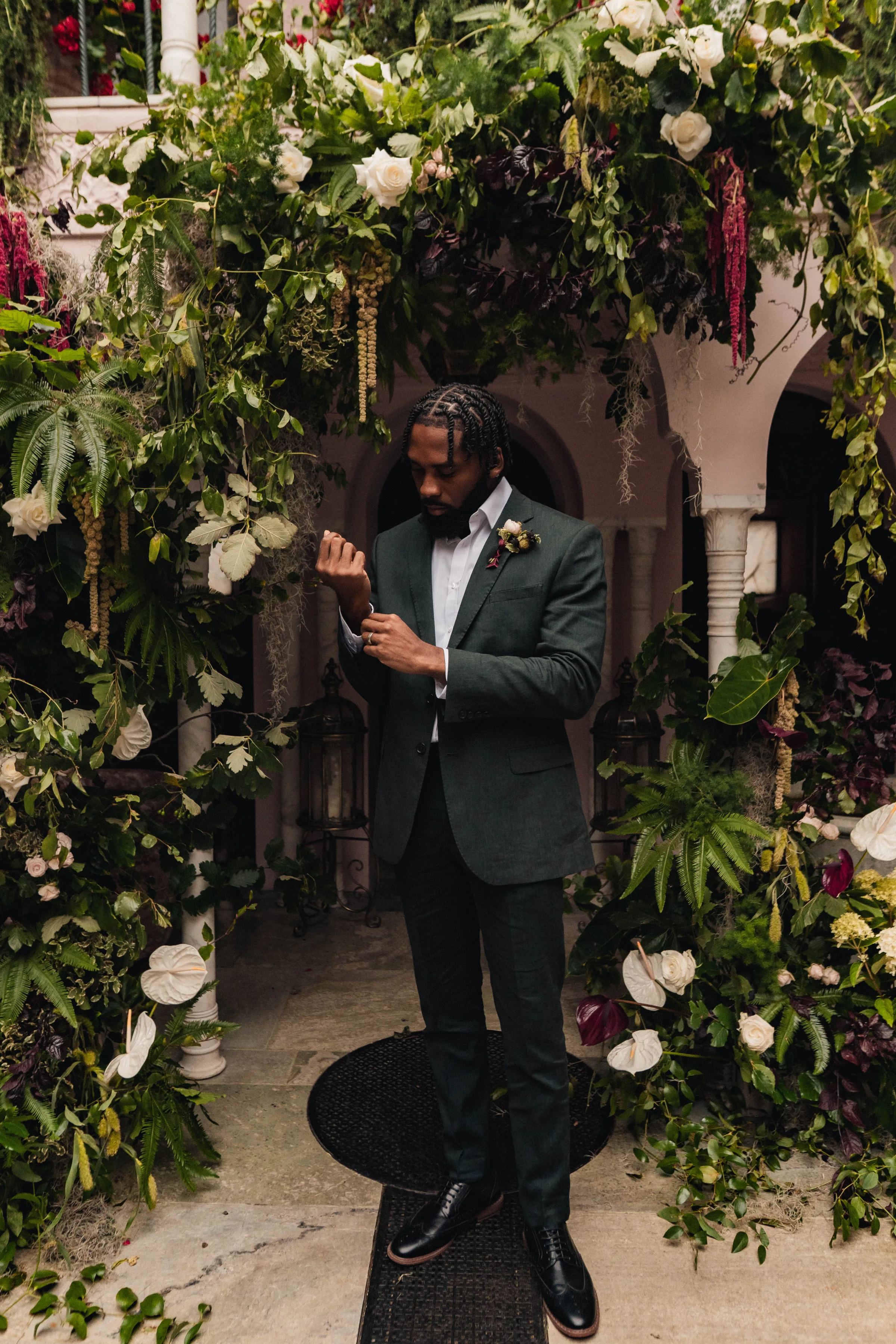 SS Port Lympne Safari Park New Orleans Wedding Editorial The Chamberlins Film Medium Format Photography London Surrey 336 - Wedding Inspiration That Celebrates The Joy, Diversity & Vibrancy of New Orleans Black model groom wearing a dark green suit standing by a backdrop of lush greenery in the form of a broken arch, featuring roses and orchids.
