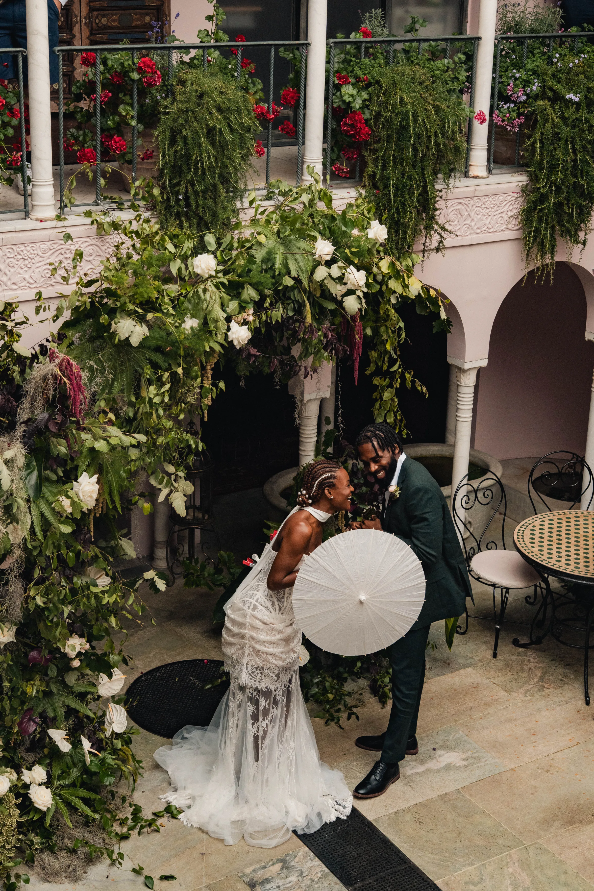 SS Port Lympne Safari Park New Orleans Wedding Editorial The Chamberlins Film Medium Format Photography London Surrey 383 1 - Wedding Inspiration That Celebrates The Joy, Diversity & Vibrancy of New Orleans Black bride and groom having a playful moment with a white parasol in the pink courtyard at Port Lympne Resort & wedding venue in Kent. They are surrounded by lush greenery, roses and large white orchids.