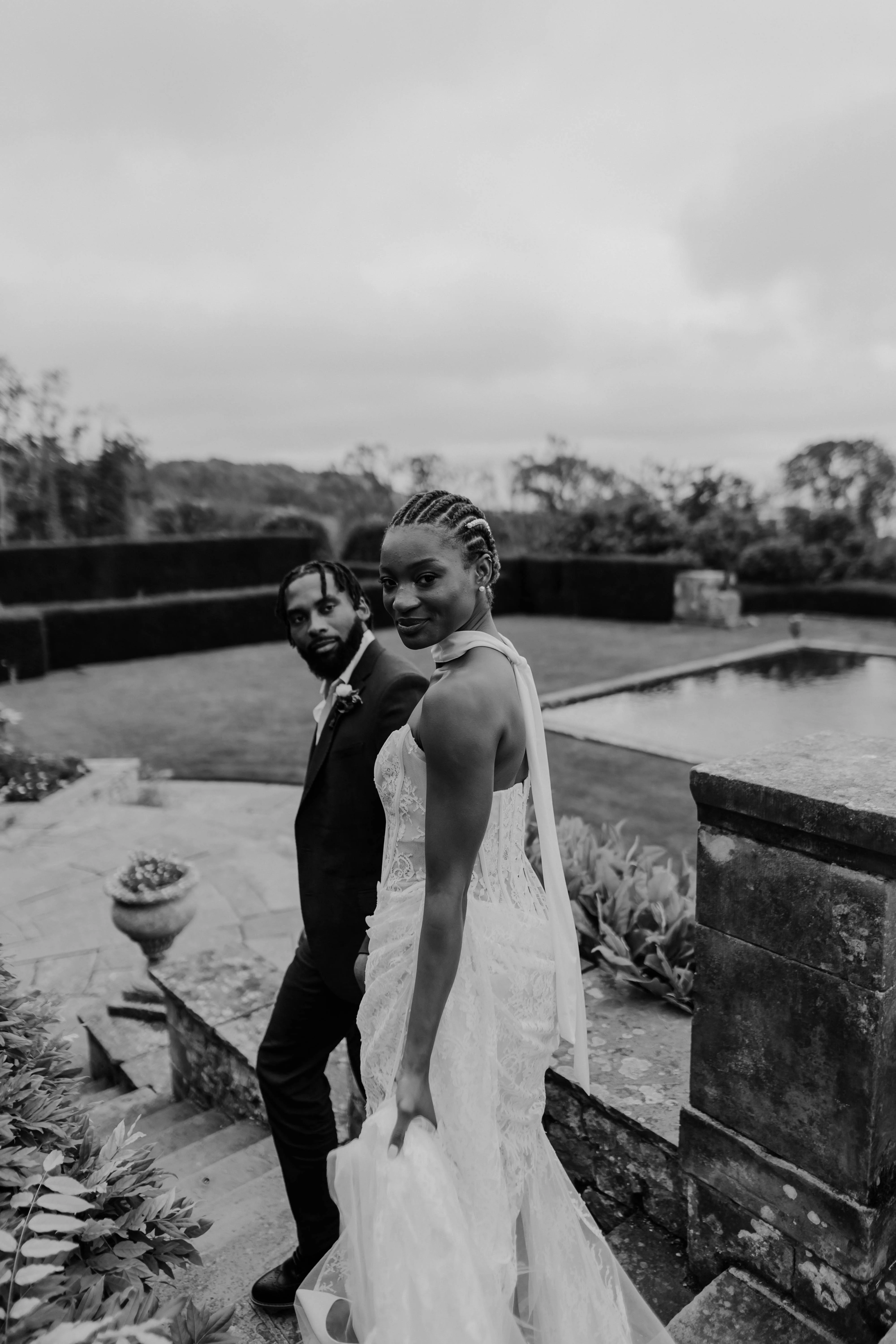 SS Port Lympne Safari Park New Orleans Wedding Editorial The Chamberlins Film Medium Format Photography London Surrey 569 - Wedding Inspiration That Celebrates The Joy, Diversity & Vibrancy of New Orleans Elegant over the shoulder shot of Black bride with pearls in her braided heir as she walks hand in hand with her groom through the grounds of Port Lympne Resort hotel and wedding venue in Kent.
