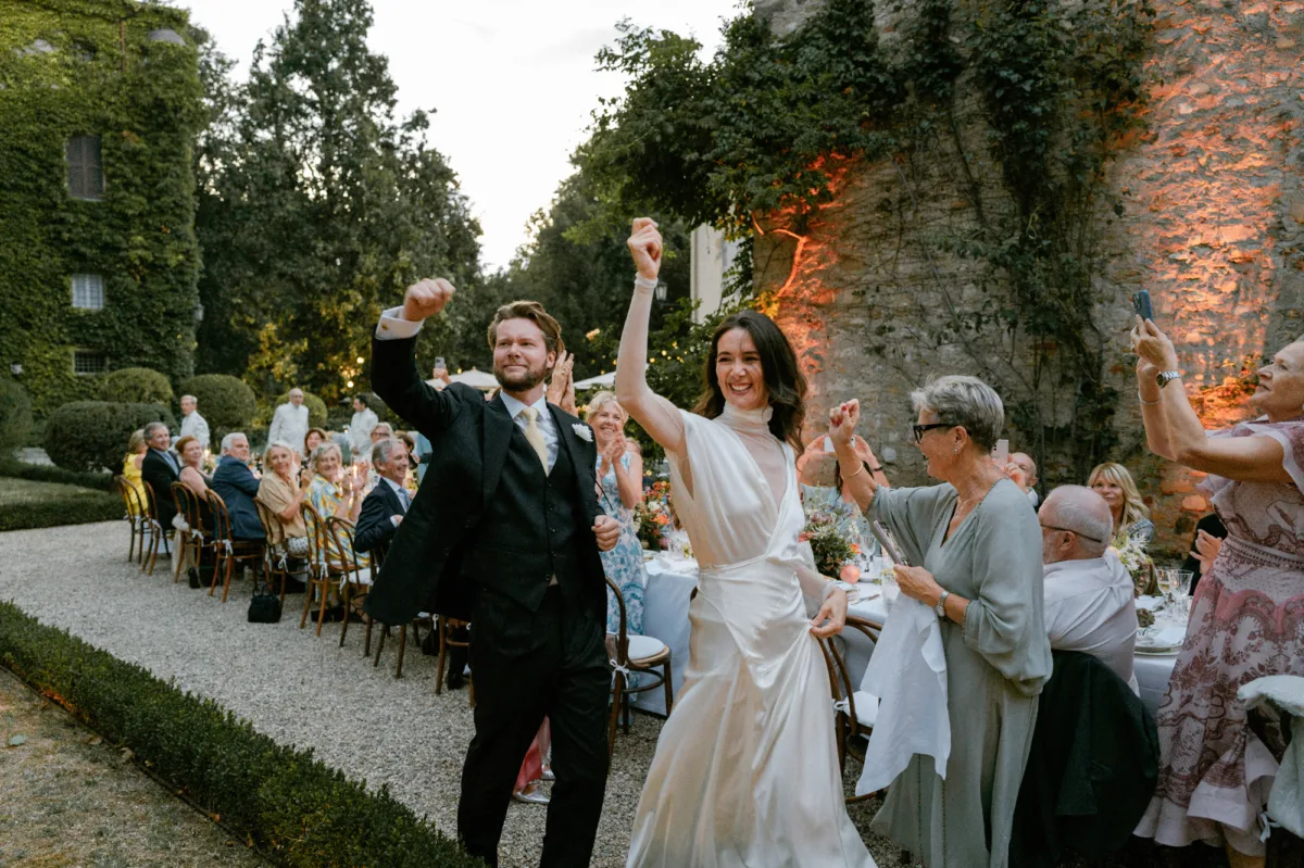 Joyous scene of bride wearing Cortana arriving to her wedding reception with her groom, hands raised in the air with excitement.