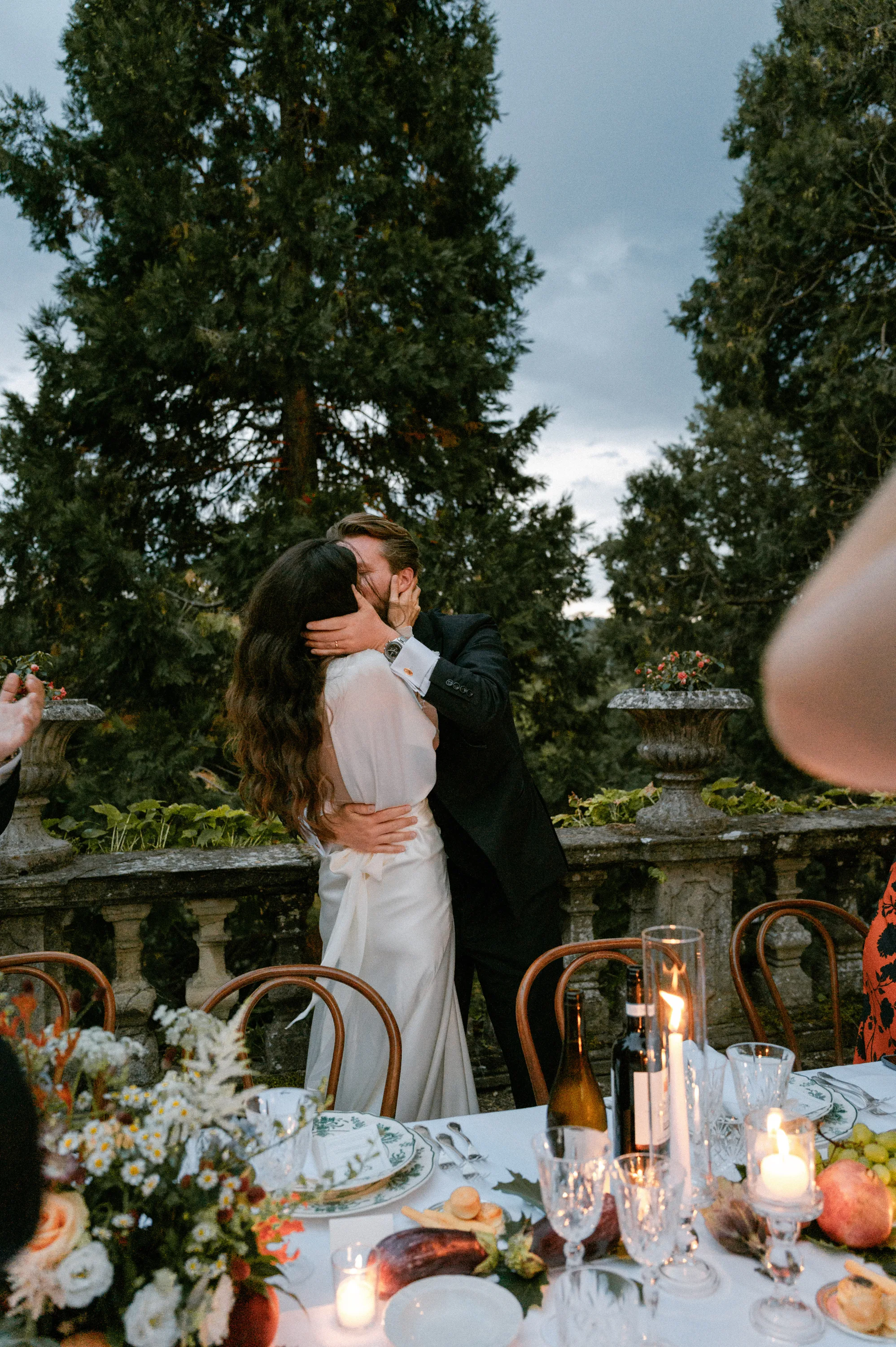 Bride wearing Cortana hugging her groom closely in a passionate embrace. They stand next to their outdoor dining table filled with candles, fruit, flowers and elegant tableware.