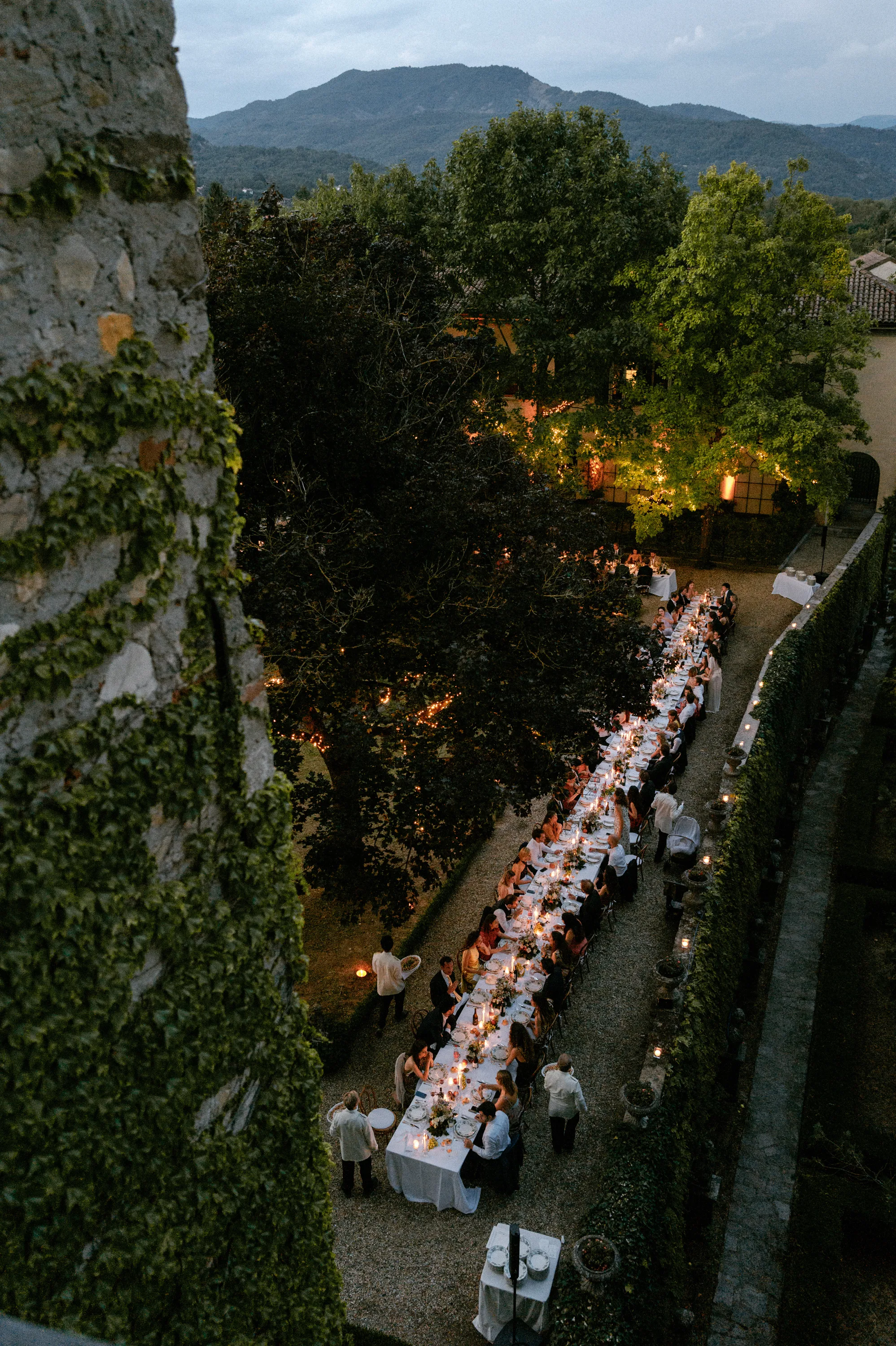 Drone shot showing the long, candlelit dining scenes of an outdoor wedding reception at a private Italian villa. Hills and mountains in the distance.