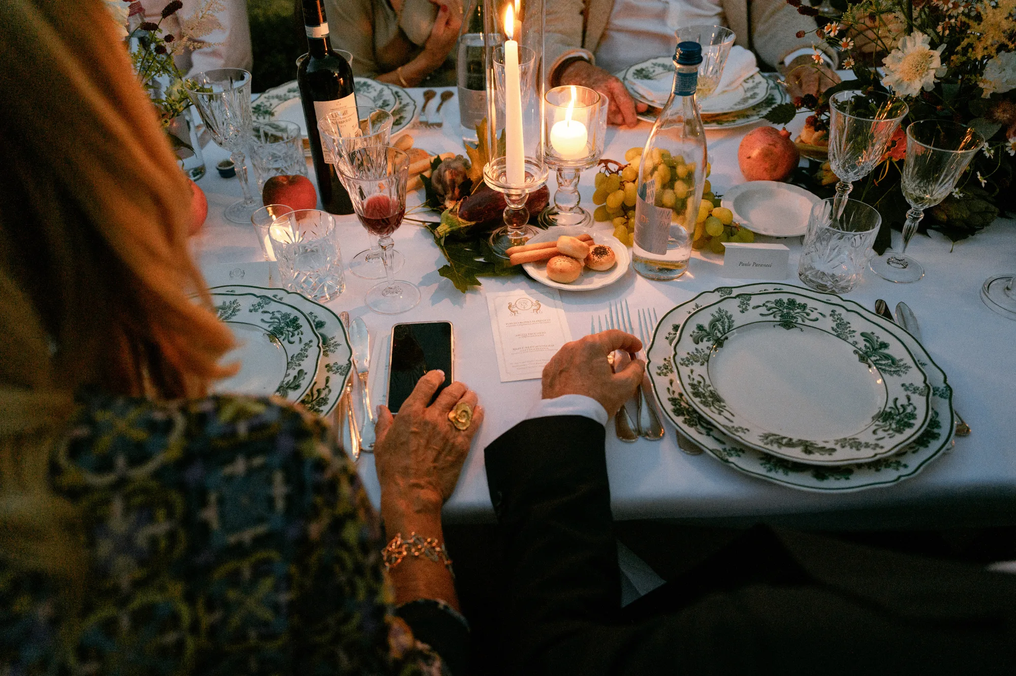 Close up shot of an intimate dining scene during an Italian outdoor wedding reception. A warm, candlelit scene with hands, fruit and glasses of wine.