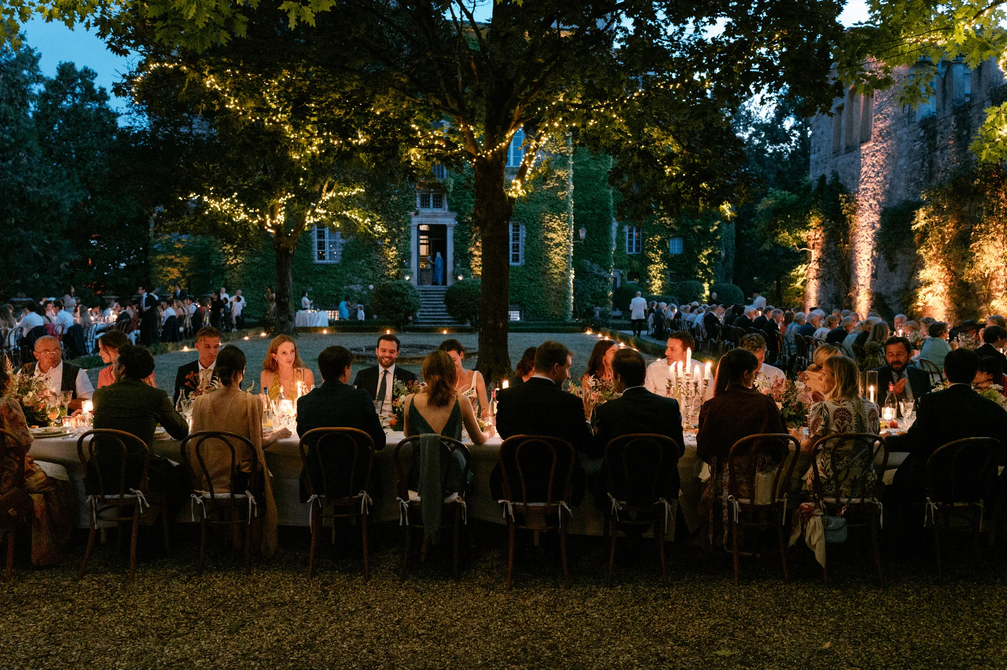 Close up shot of an intimate outdoor evening wedding dinner with guests dining under trees in a candlelit scene.