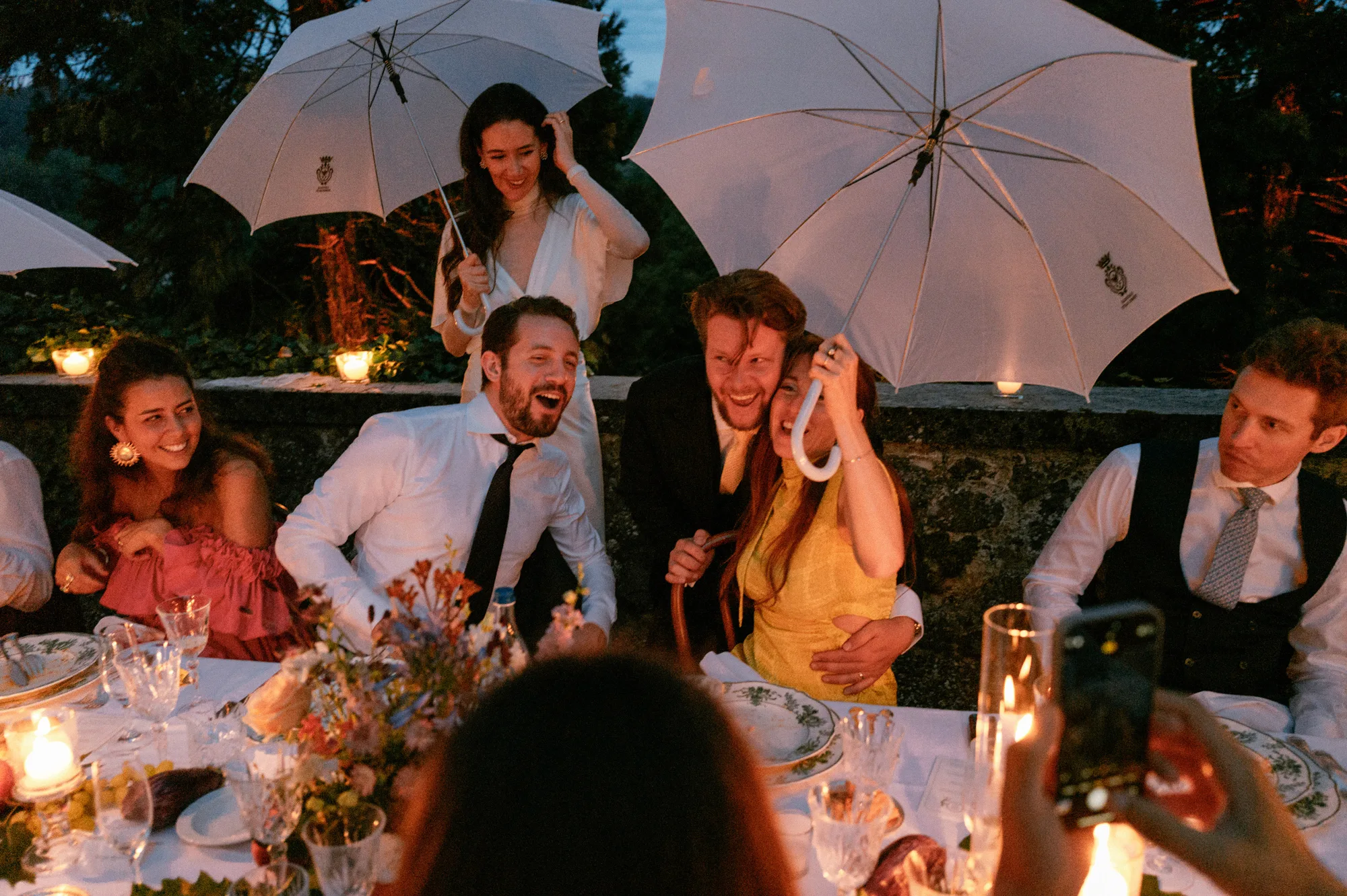 Close up shot of an intimate outdoor evening wedding dinner with the bride and guests holding white parasols up to protect them from a light rain shower.