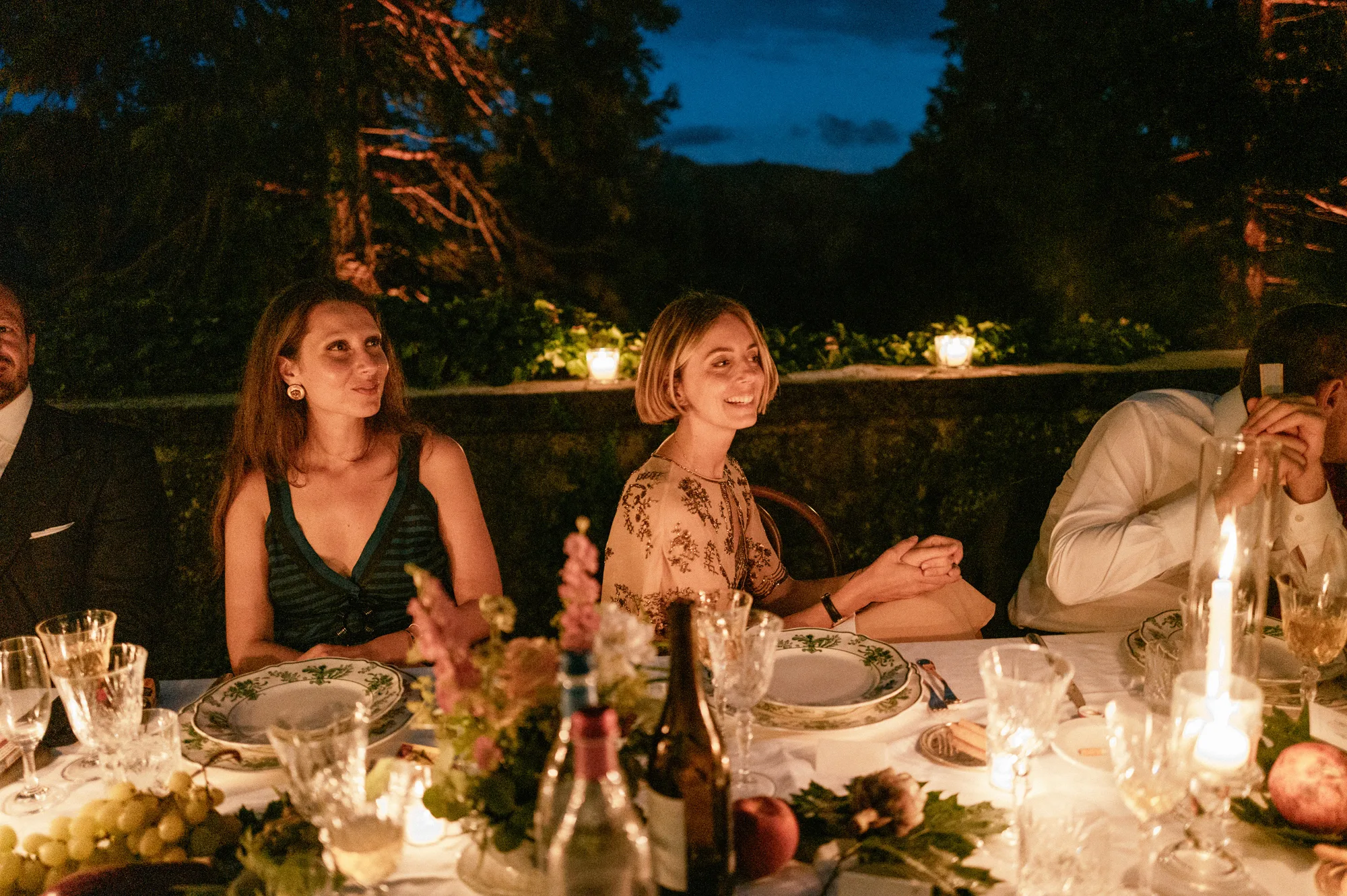 Close up shot of an intimate outdoor evening wedding dinner with guests dining under trees in a candlelit scene.