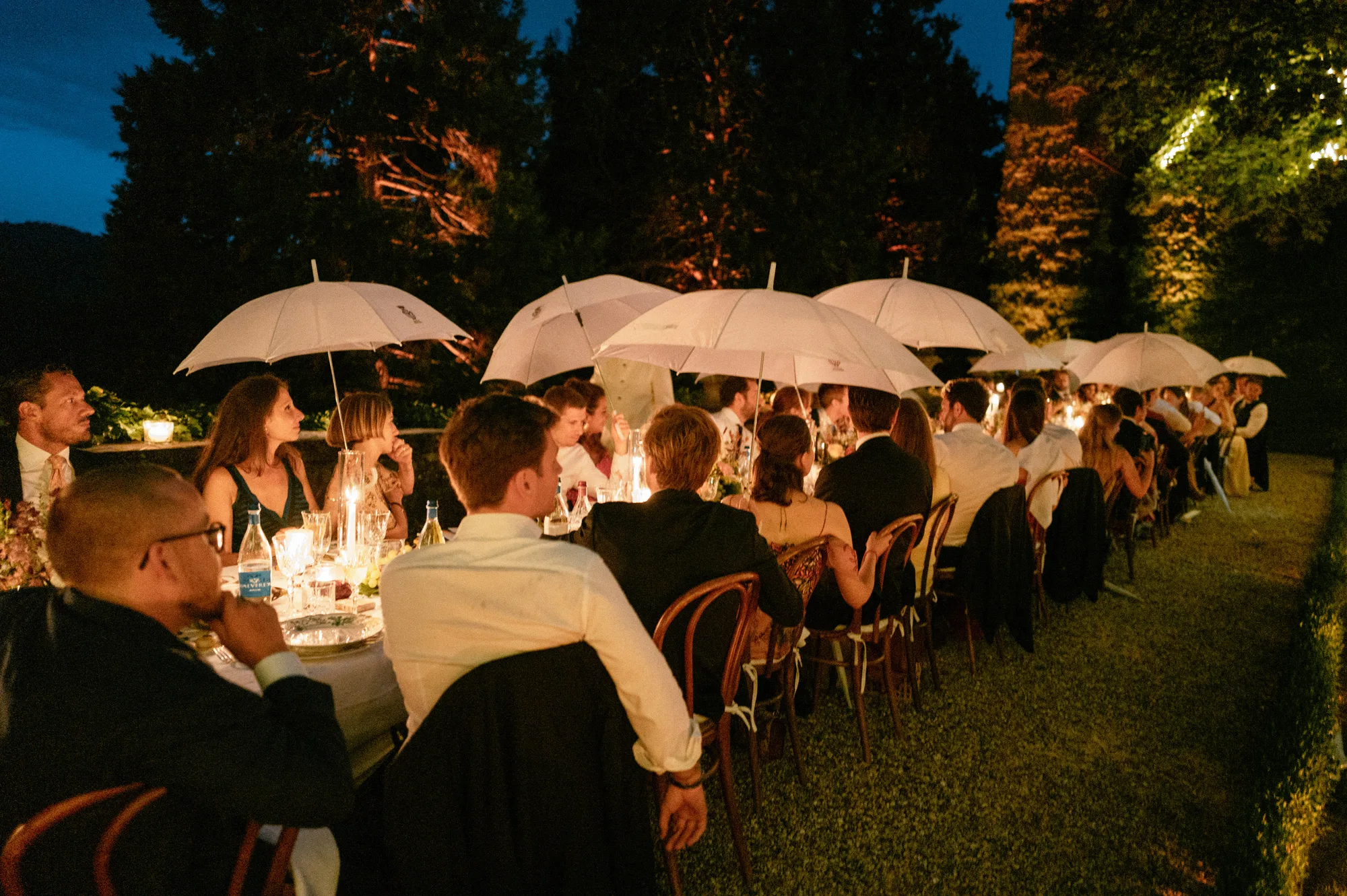 Close up shot of an intimate outdoor evening wedding dinner with the bride and guests holding white parasols up to protect them from a light rain shower.
