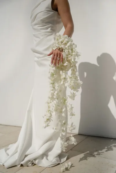 Bride in a fitted floral-applique gown holding flowers with soft shadows on the wall, a wedding portrait