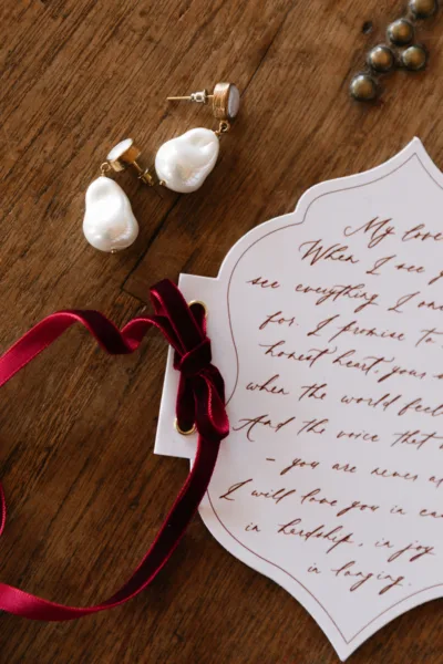 Wedding stationery flat lay with a handwritten letter, pearl earrings and burgundy ribbon, styling by Zahra Studio Events