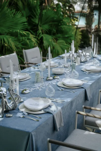 Outdoor wedding reception table with dusty-blue linens and candles among palm trees, design by Zahra Studio Events
