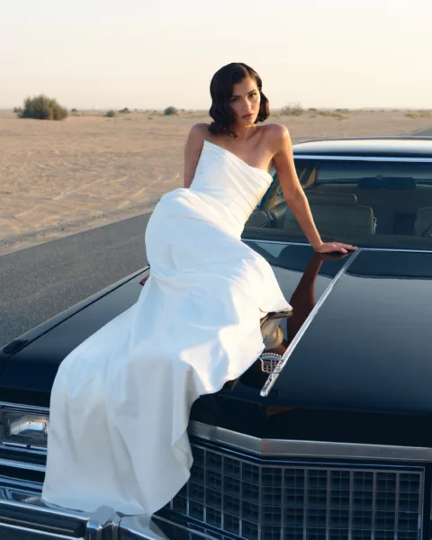 Bride in a white gown beside a black vintage car in a desert landscape during a destination wedding