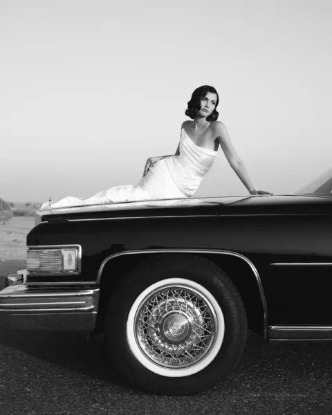 Black and white portrait of a bride in a white gown leaning on a vintage car at a destination wedding