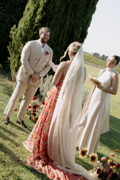 Couple at an outdoor Tuscan wedding ceremony with an officiant and sunflowers, the bride in a red floral gown