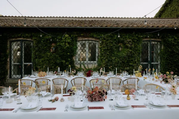 Long outdoor wedding reception table set against an ivy-covered Tuscan villa with string lights, design by Zahra Studio Events