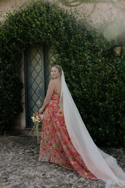 Bride in a red floral-print gown with a long train beside an ivy-covered archway at a Tuscan villa wedding