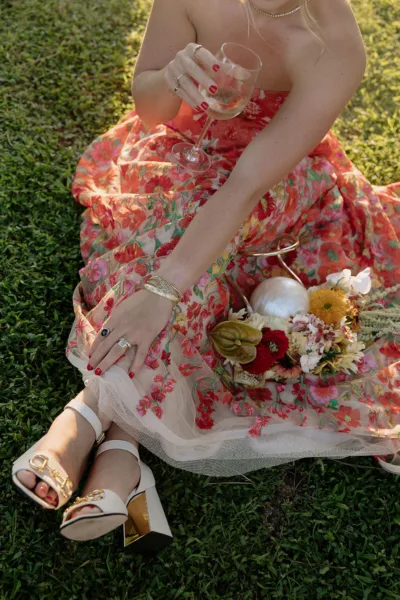 Bride seated on the grass in a red floral-print gown with a bouquet and gold heels at a Tuscan wedding