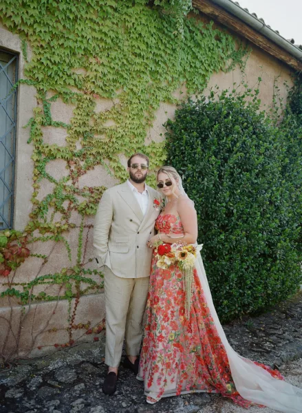 Newlyweds posing by a hedge, the groom in a cream suit and the bride in a red floral gown at a Tuscan villa wedding