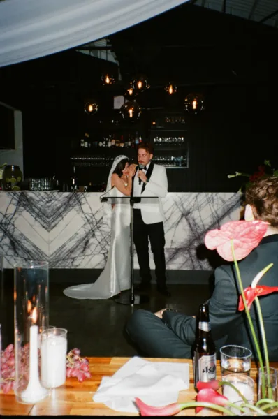 Groom giving a speech beside the bride at their candlelit wedding reception in a candid documentary-style photograph.