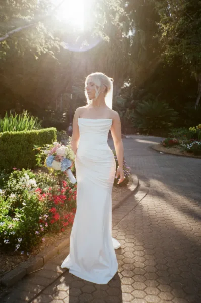 Editorial bridal portrait in a sunlit garden, with the bride carrying a colourful bouquet and wearing a modern strapless wedding dress.