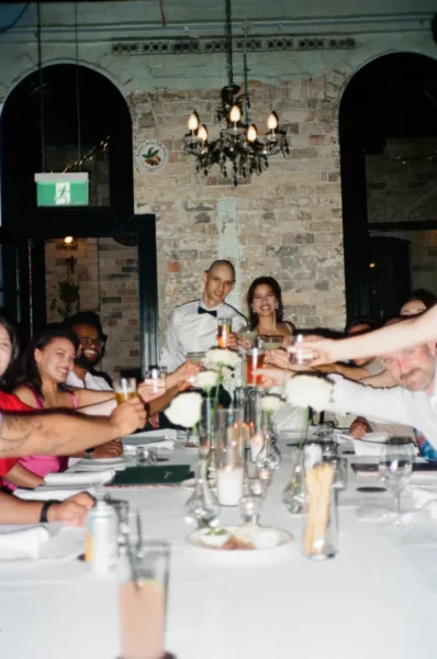 Newlyweds and their guests raising glasses around a long candlelit reception table in a candid wedding photograph.