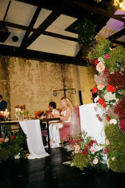 Bride seated at her wedding reception beneath a dramatic installation of red, pink and green flowers.