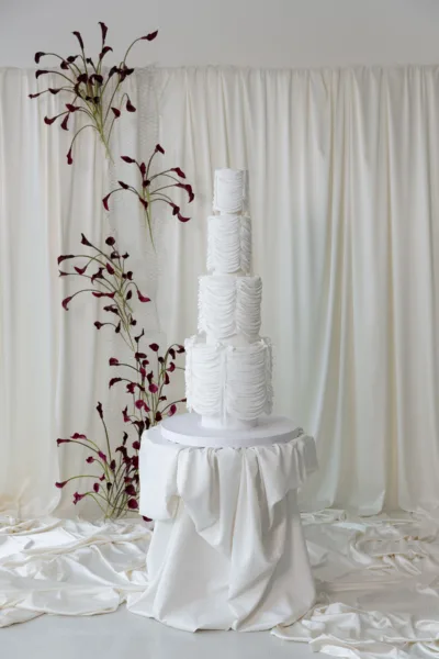 White tiered wedding cake accented with dark calla lily branches against white drapery, design by Zahra Studio Events
