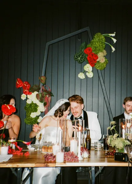 Bride and groom sharing a candid moment at their wedding reception beneath bold red and white floral arrangements.