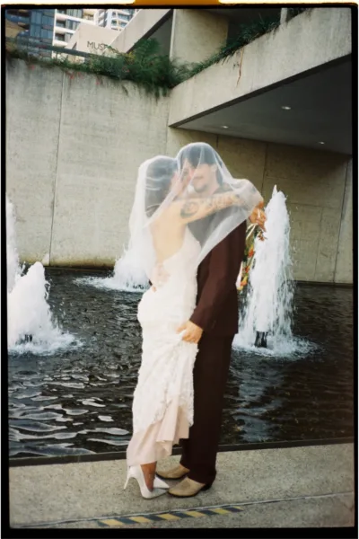 Full-length portrait of a bride and groom kissing beneath her veil beside a contemporary water feature.