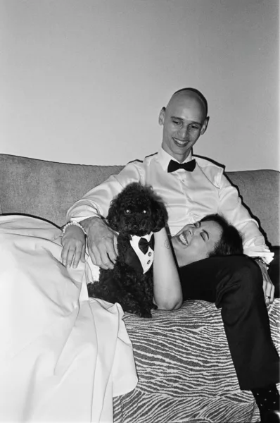 Groom seated with a small black dog wearing a bow tie in a candid black-and-white wedding photograph.