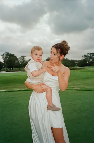 Bride holding her baby on the lawn during an outdoor wedding, captured in candid 35mm film photography.