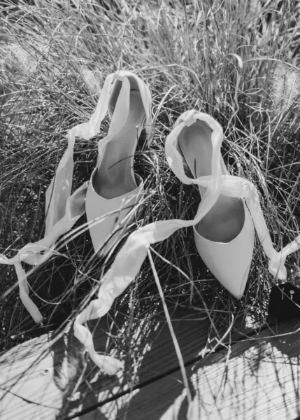 White bridal shoes with ankle ribbons arranged in long grass in a black-and-white wedding photograph.