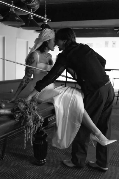 Bride wearing a bandana-style veil beside a pool table in a candid black-and-white wedding photograph.