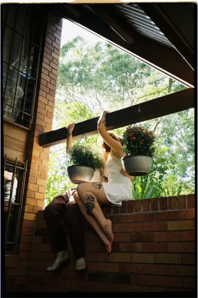 Newlyweds sitting together beneath hanging plants in a playful editorial wedding portrait captured on film.