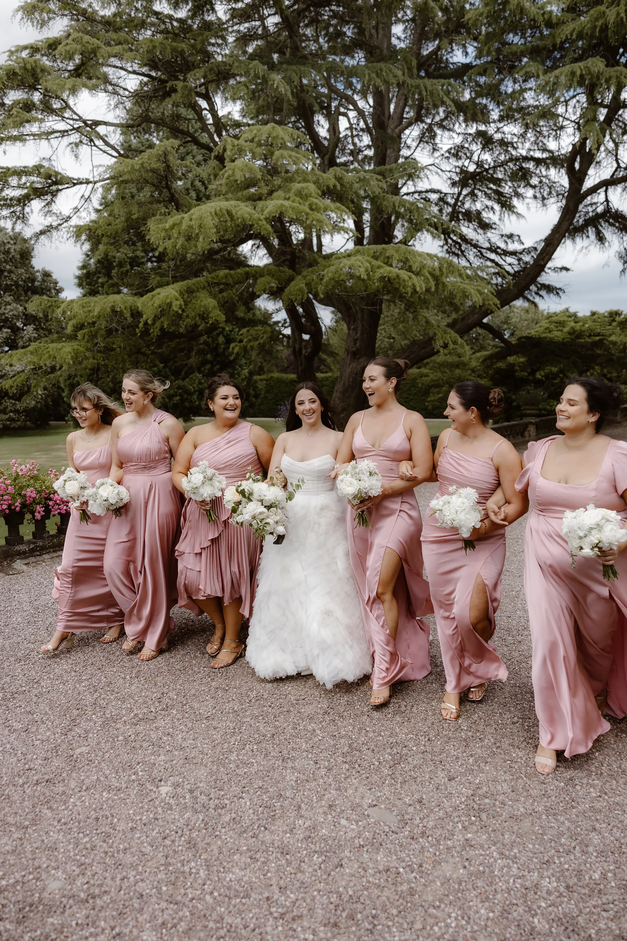 Capesthorne Hall Summer Wedding Jessica Sharpe Wedding Planner Jono Symonds Love My Dress.53 - Sophie & Dimitri's Beautiful, Relaxed & Elegant Wedding Weekend at Capesthorne Hall in Cheshire Bride and her bridesmaids looking feminine and elegant as they wander arm in arm the ground of Capesthorne Hall in Cheshire. The bridesmaids wear pink dresses by Amaze. The bride wears a Watters dress. They all carry bouquets of white flowers.