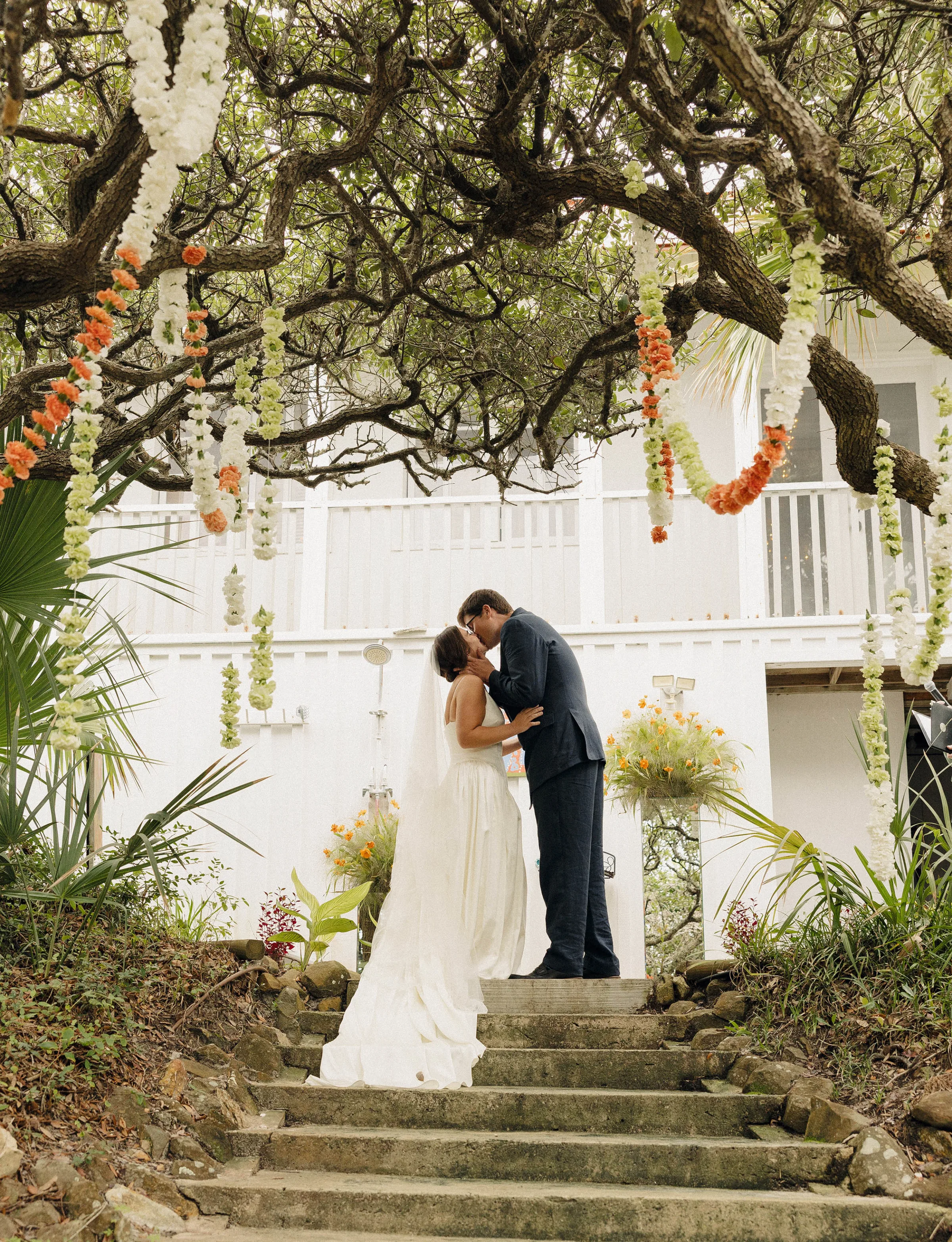 Danielle Frankel Bride Leslie DeAnne Photography.49 - Olivia in Danielle Frankel for her Intimate, Modern & Non-Traditional Wedding Bride and groom kissing one another on the top of a set of stone stairs. Above them hang orange and green garlands from the trees. She wears a Danielle Frankel gown and they are surrounded by orange and green flowers.