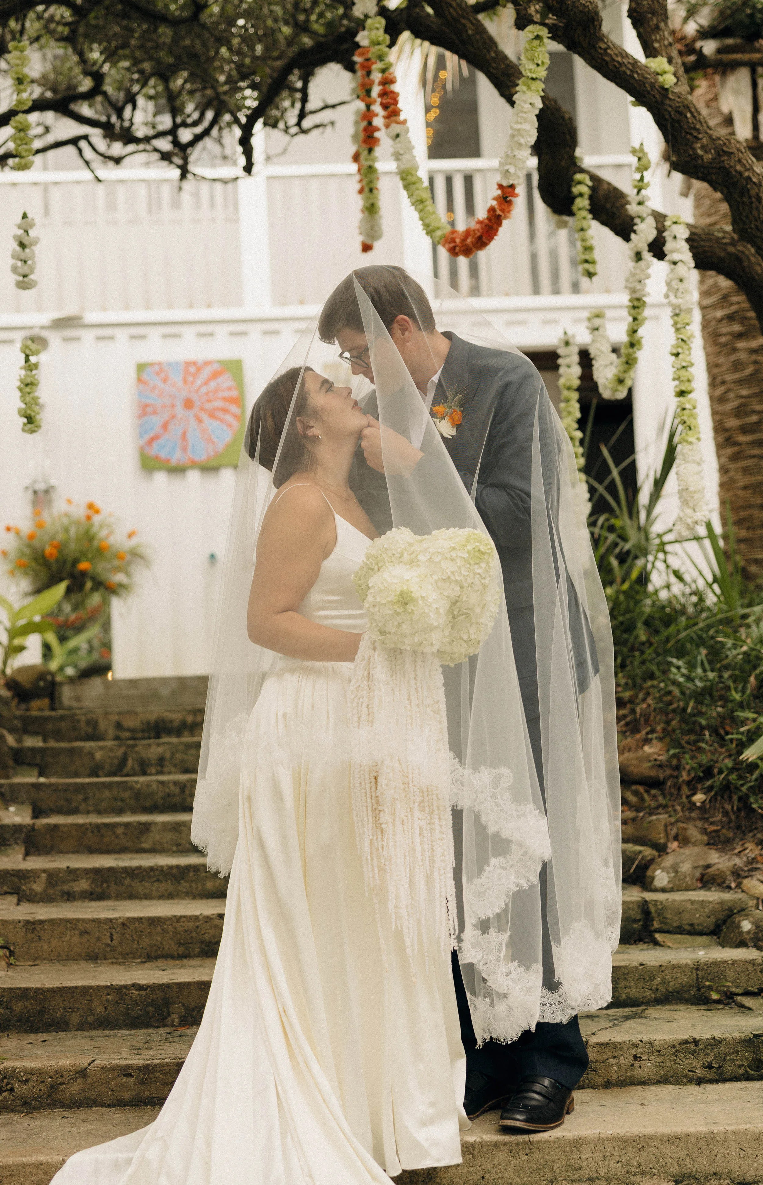 Danielle Frankel Bride Leslie DeAnne Photography.56 - Olivia in Danielle Frankel for her Intimate, Modern & Non-Traditional Wedding Bride and groom kissing one another on the top of a set of stone stairs. Above them hang orange and green garlands from the trees. She wears a Danielle Frankel gown and they are surrounded by orange and green flowers.