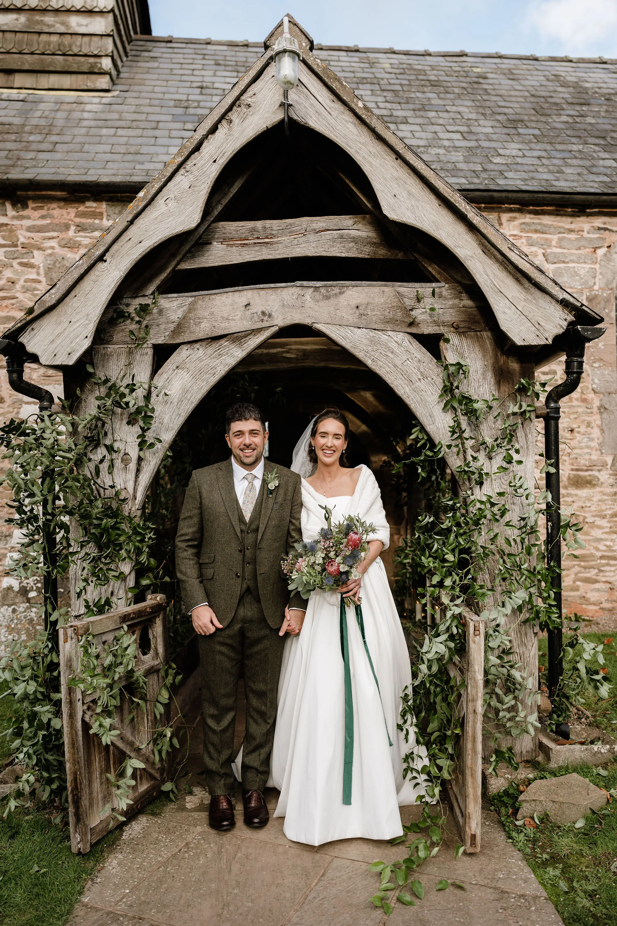 Bride and groom standing beneath a weathered wooden church lychgate draped in trailing foliage, bride holding a wildflower bouquet with thistles, protea and eucalyptus with dark green ribbon streamers.