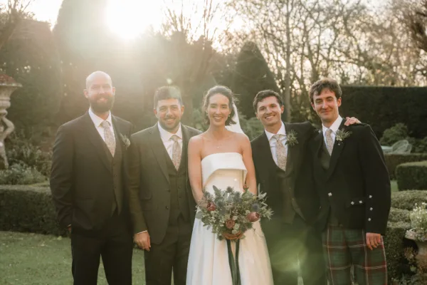 Bride holding a thistle and protea bouquet standing with four groomsmen in a formal garden at golden hour, one wearing a tartan kilt, crystal chandeliers and topiary hedging in the background.