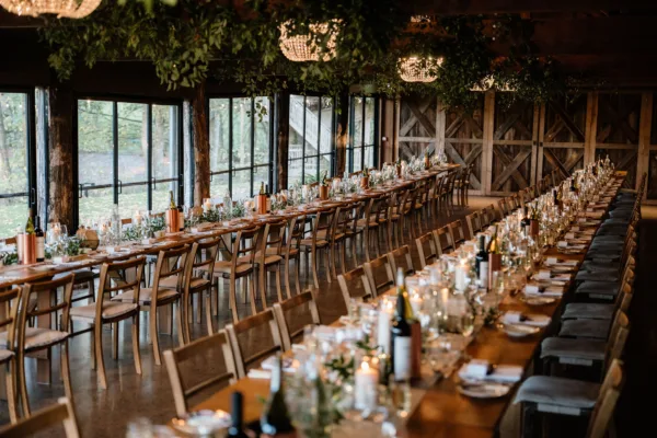 Long oak feasting tables set for a wedding reception inside a barn venue, with abundant greenery ceiling installation woven around crystal chandeliers, candlelight and copper vessel centrepieces.