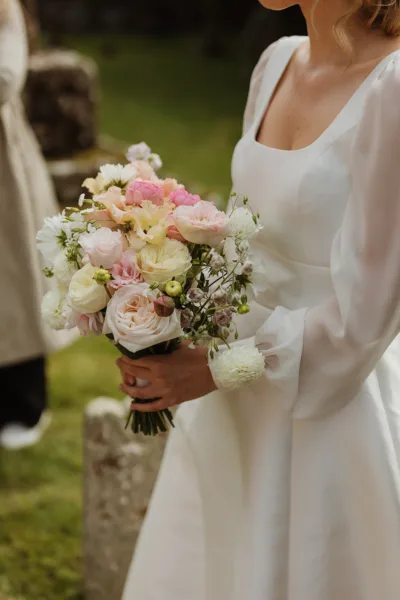 Close-up of a bride's hands holding a soft, abundant bouquet of blush garden roses, peach ranunculus, cream spray roses, pink lisianthus and white scabiosa against a white long-sleeved wedding dress.