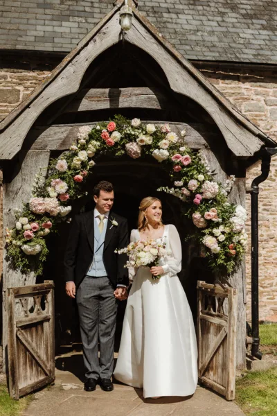 Bride and groom walking hand in hand through a stone church lychgate framed by a full circular floral arch of blush, coral and ivory roses, peonies and dahlias with deep green foliage.