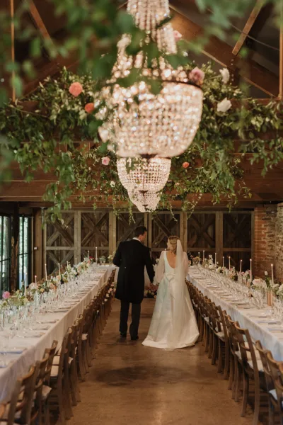 Bride and groom walking hand in hand down the aisle of a barn wedding reception, viewed from behind, with long candlelit feasting tables either side, flowers and greenery ceiling installation above, and crystal chandeliers glowing overhead.