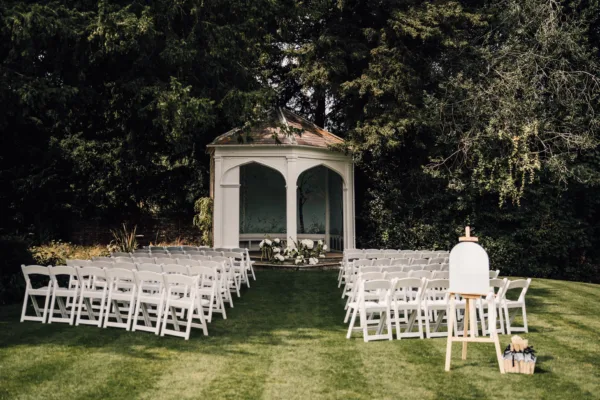 Outdoor wedding ceremony setup with rows of white chairs on a lawn and a white arched gazebo