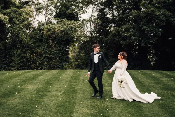 Bride in a flowing dress and groom walking together across a green lawn at a wedding venue