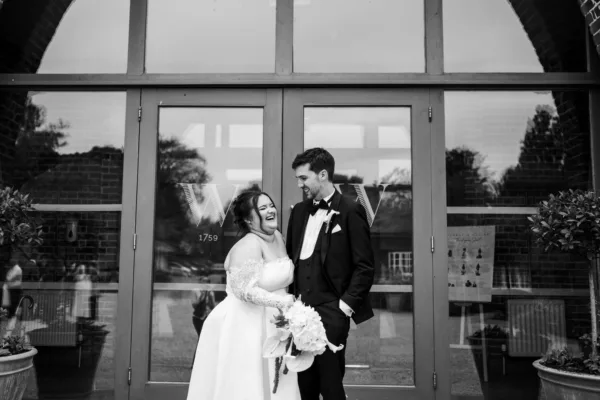 Black and white photo of a groom standing at large glass doors inside a modern wedding venue