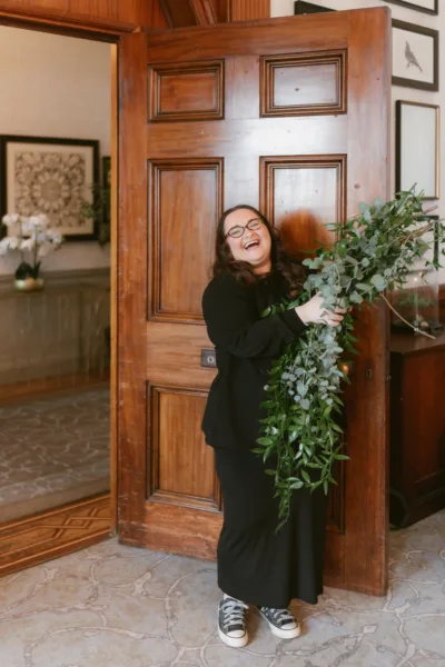Wedding planner Lauren Batten laughing while arranging a large greenery installation at a venue entrance