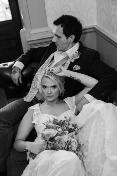 Black and white portrait of a bride in a lace-detail dress seated and holding a bouquet
